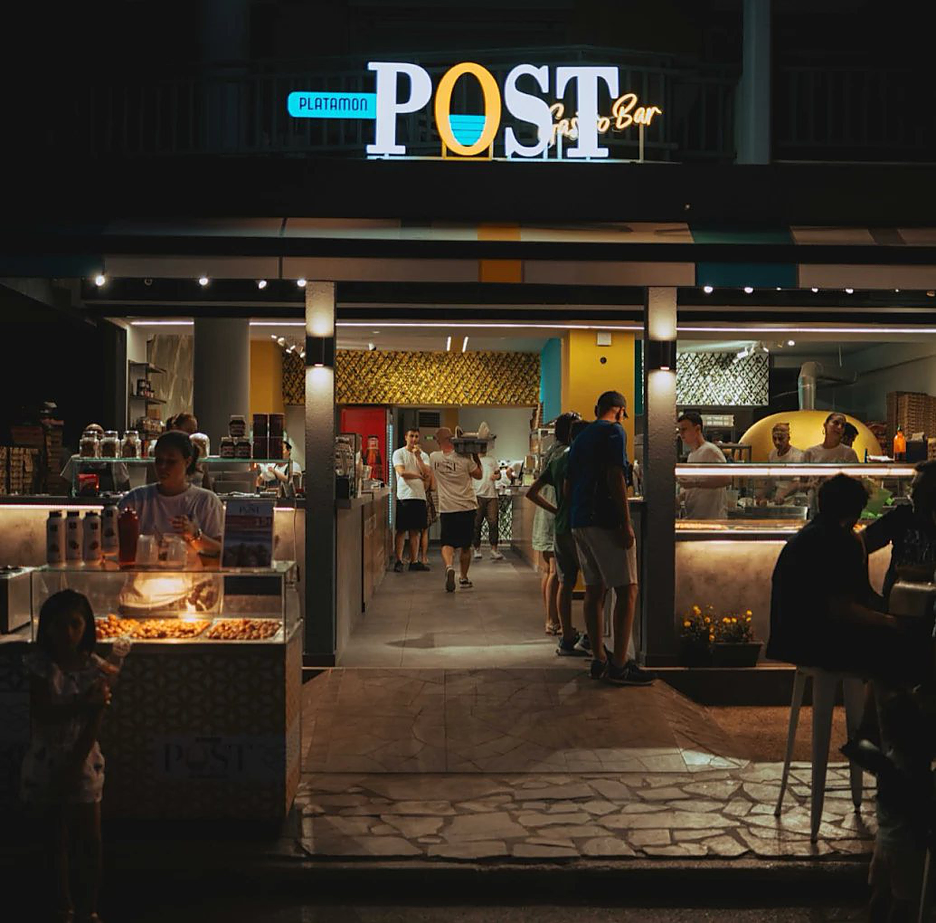 Η φωτισμένη είσοδος του Platamon Post το βράδυ, με κόσμο στον πάγκο